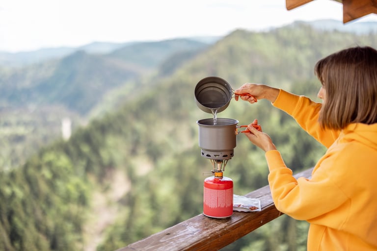 Young woman prepares food in special dishware for hiking and portable gas burner, while traveling in mountains on a summertime. Concept of camping equipment and traveling on nature