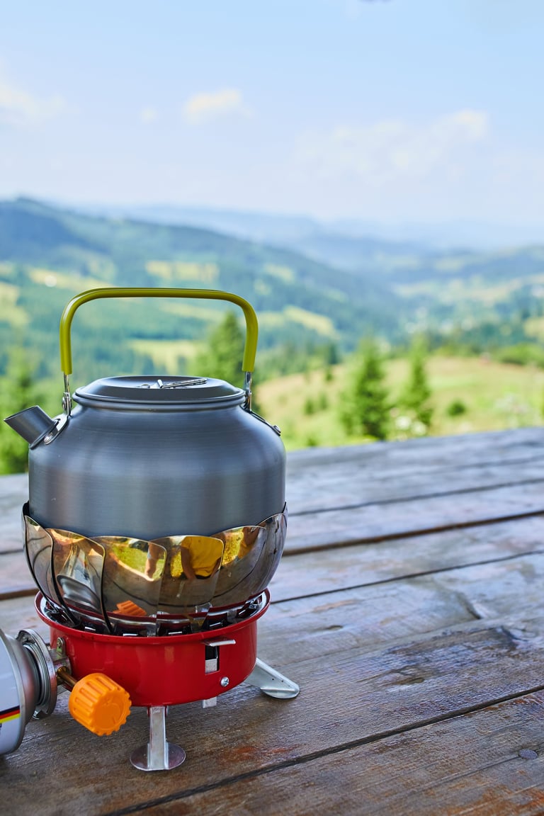 A kettle sits on a red camping stove, surrounded by a breathtaking view of green mountains and trees, capturing a peaceful outdoor moment in nature.