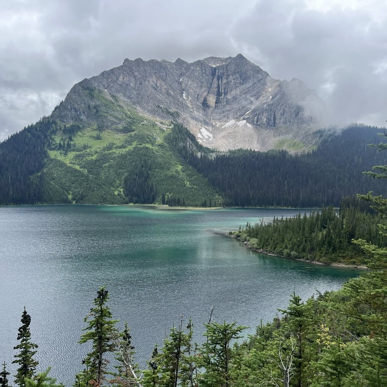 Alpine lake with turquoise water surrounded by evergreen forest and rocky mountain peak under cloudy sky