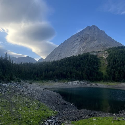 Mountain lake surrounded by coniferous forest with steep rocky peak and cloudy blue sky