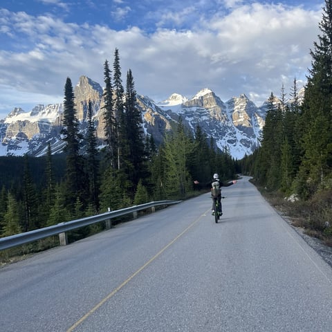 Cyclist riding on a mountain road with snow-capped peaks and evergreen forests in the background