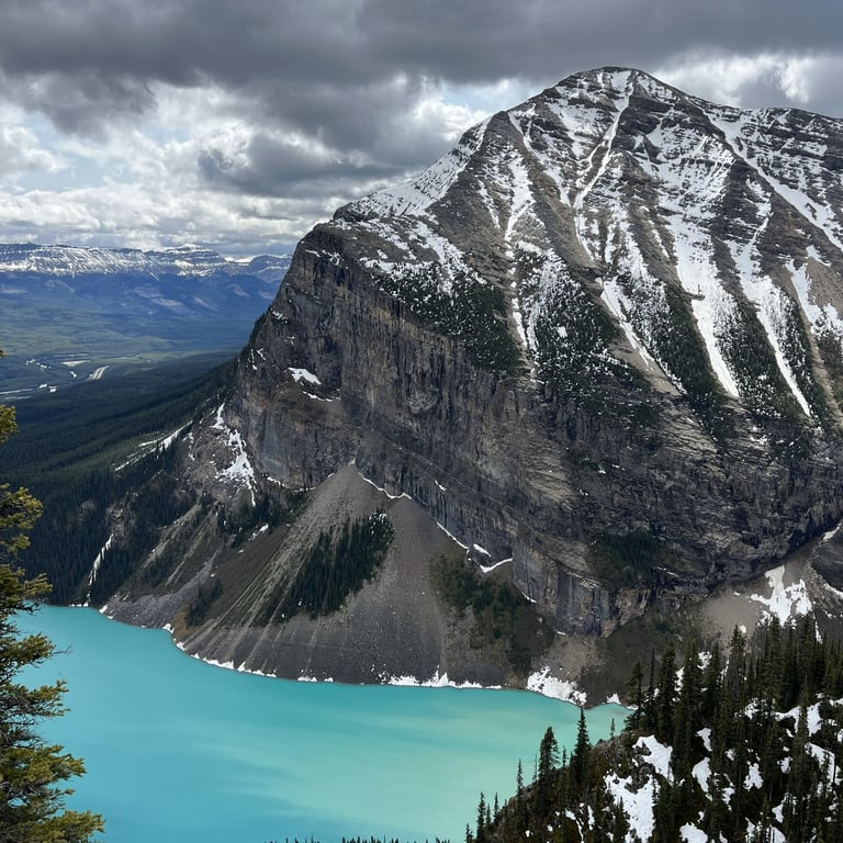 Snow-capped mountain peak overlooks a brilliant turquoise glacial lake surrounded by evergreen forest and distant valley landscape