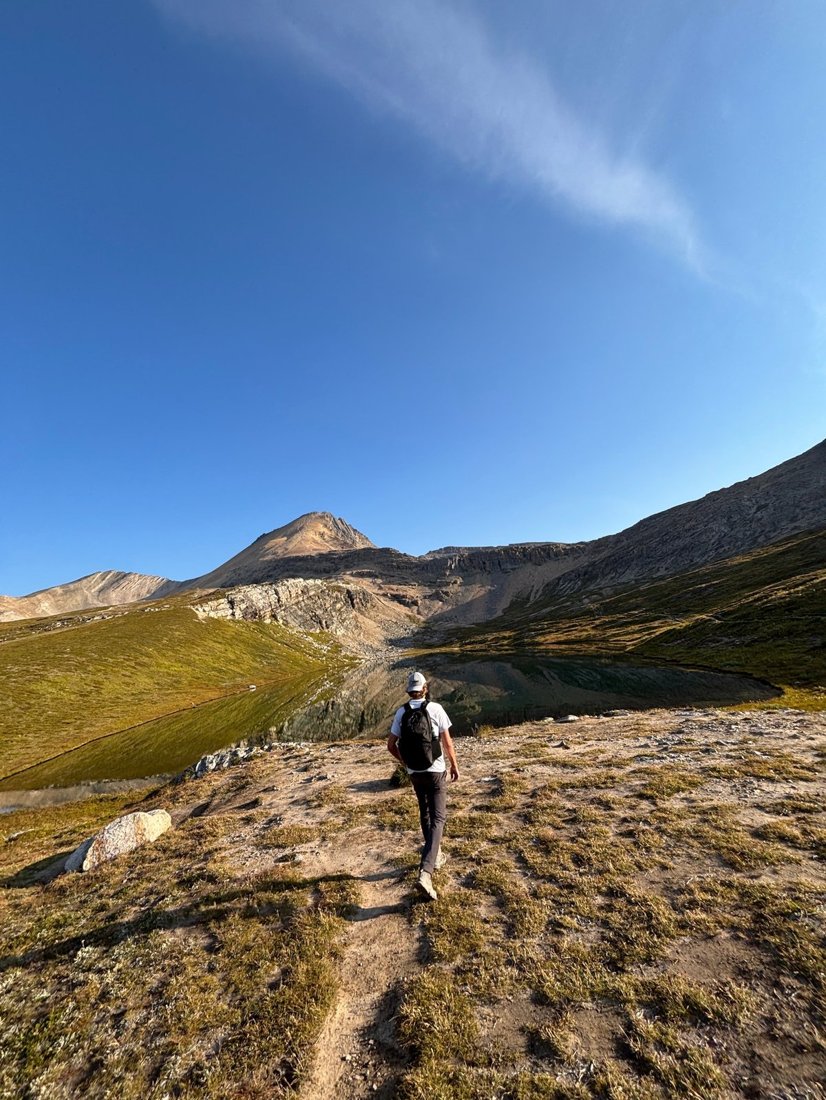 Hiker with backpack standing on rocky terrain by a mountain lake with peaked mountains in the background under blue sky