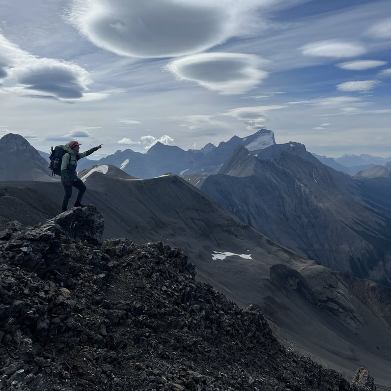 Hiker standing on rocky mountain peak with arm raised, overlooking vast alpine landscape with distant snow-capped peaks and blue sky