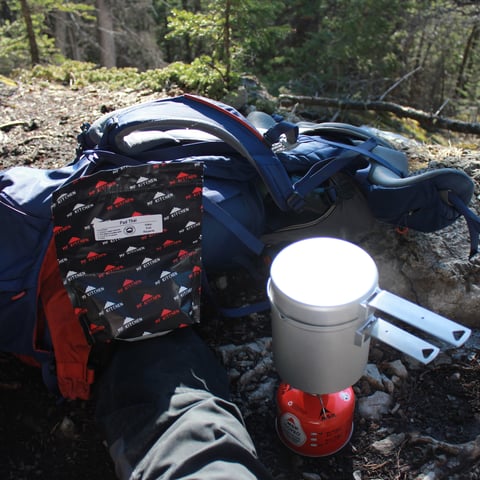 Camping gear laid out on forest ground including a blue backpack, red thermos, and clothing with trees in background