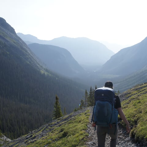 Hiker with backpack walking on mountain trail overlooking forested valley and distant peaks