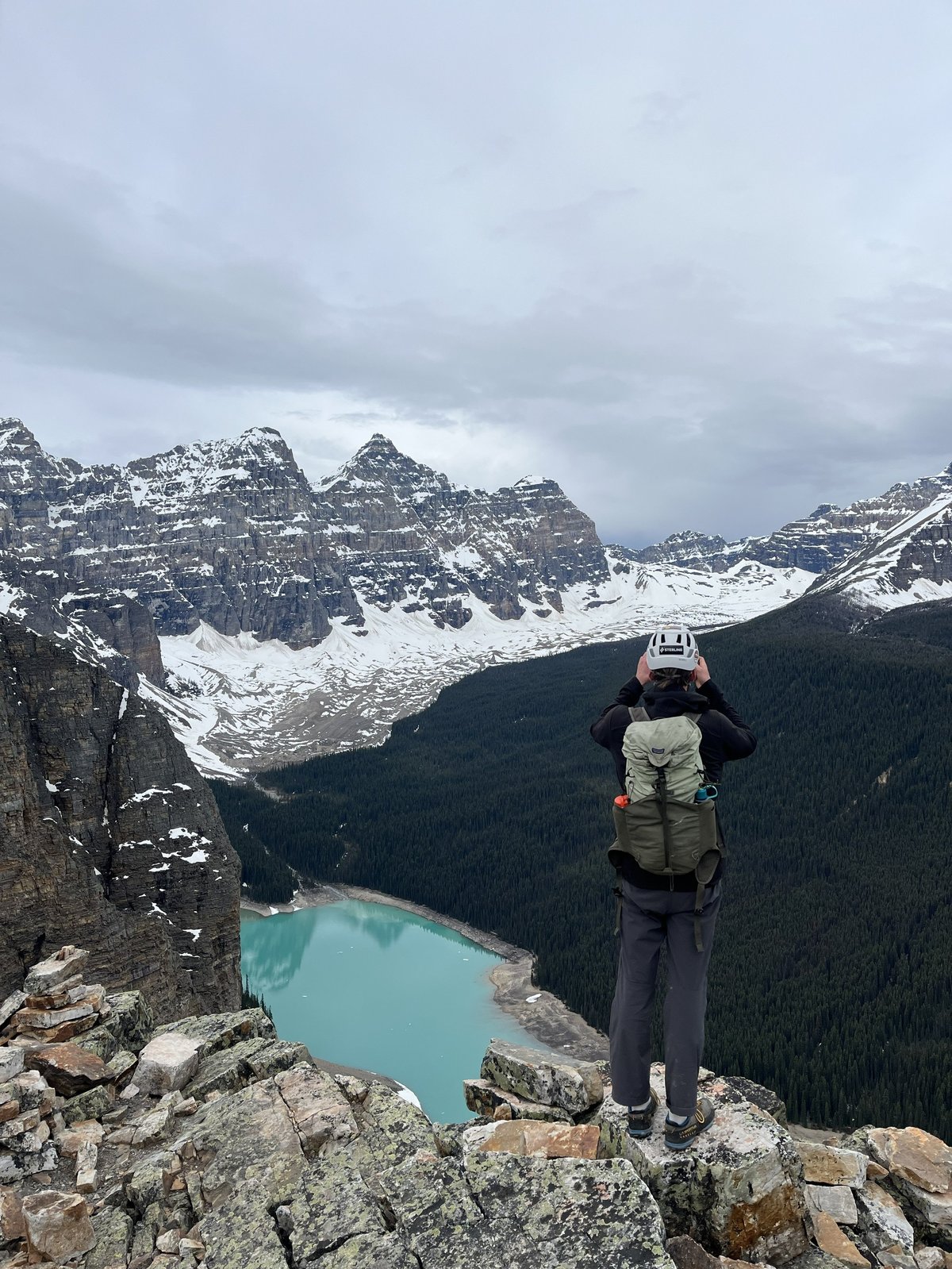 Hiker with backpack standing on rocky outcrop overlooking turquoise glacial lake surrounded by snow-capped mountains and evergreen forest
