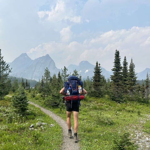 Backpacker jogging on mountain trail with rocky peaks and evergreen forest in background under blue sky