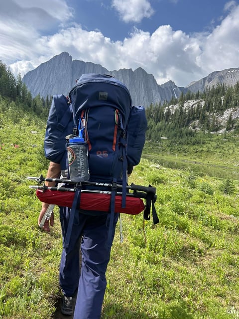Backpacker with large blue hiking backpack carrying red sleeping pad in mountain valley with granite peaks and evergreens