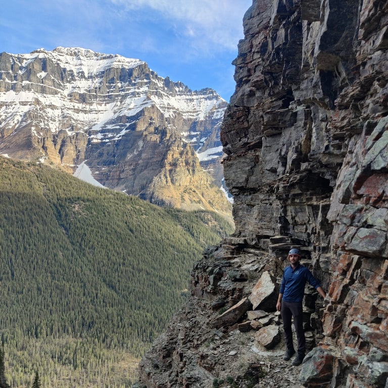 Hiker in blue jacket walking along narrow mountain trail carved into rocky cliff face with forested valley and snow-capped peaks below