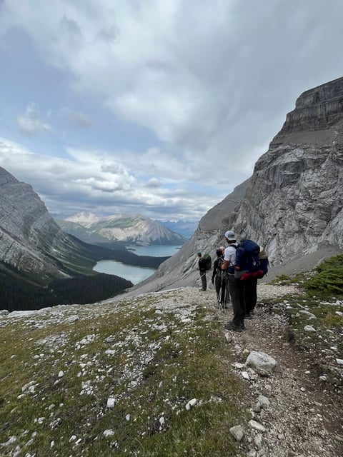 Hikers on mountain trail overlooking alpine lake surrounded by rocky peaks and forested slopes
