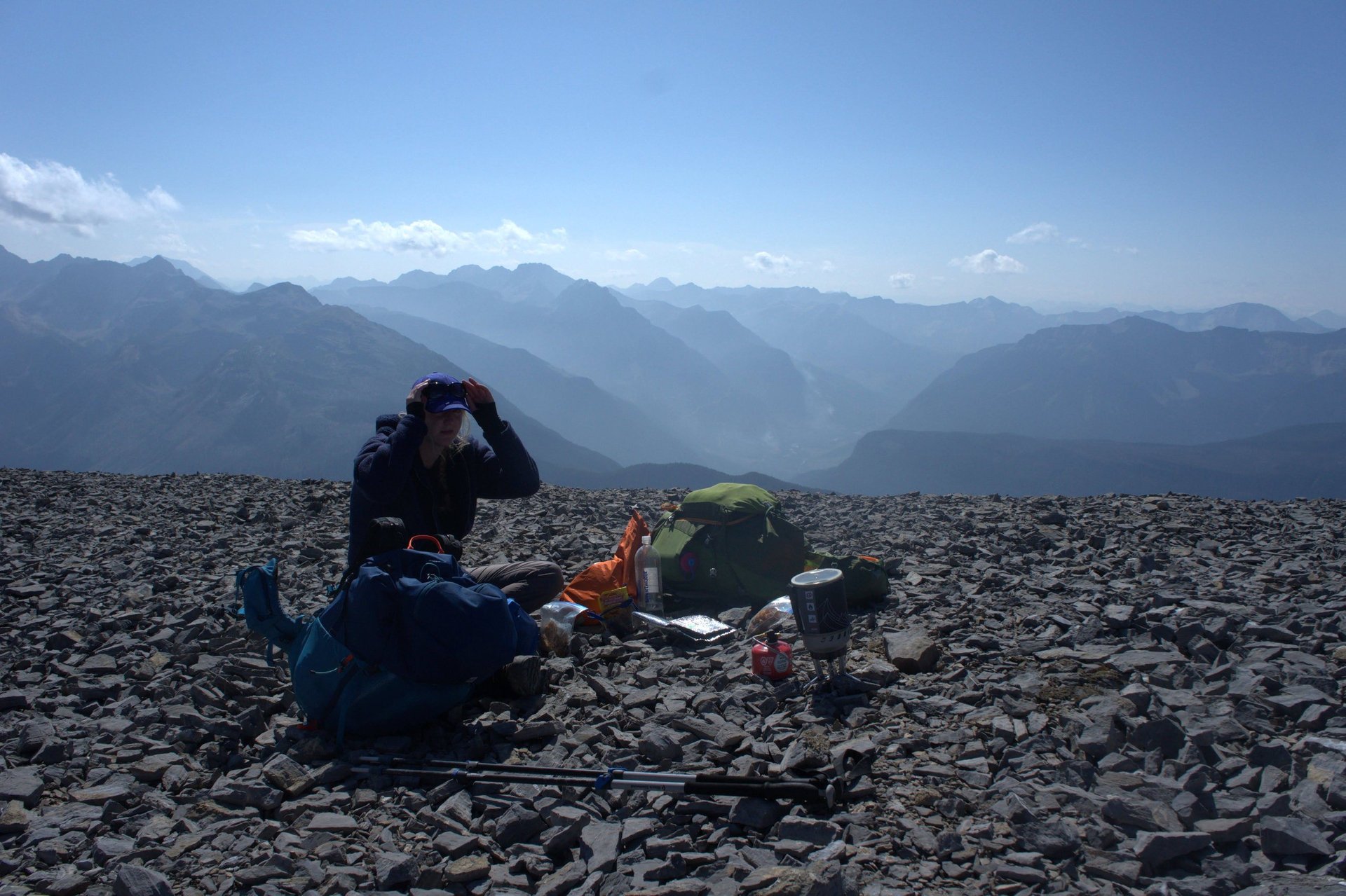 Hiker resting on rocky mountain slope with green tent and backpack, mountain range visible in misty background