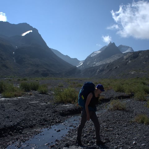 Backpacker standing in mountain valley with snow-capped peaks, blue sky, and sparse vegetation in background