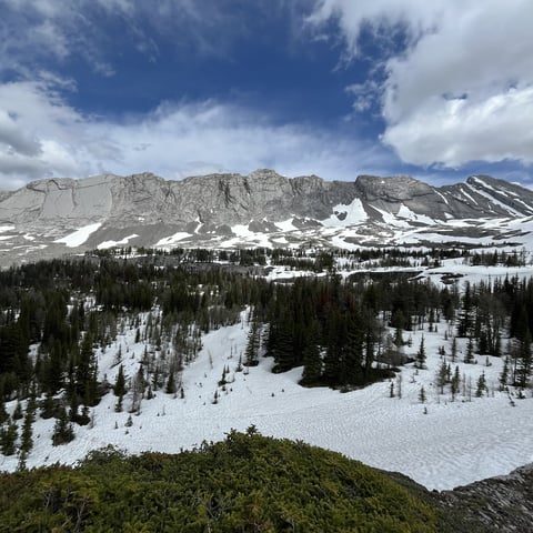 Alpine landscape with snow-covered peaks, evergreen forest, and a frozen lake under blue sky with white clouds