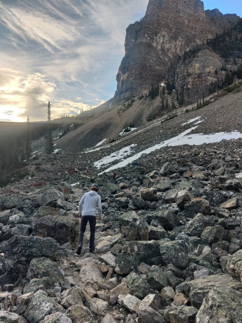 Hiker in white shirt hiking across rocky alpine terrain with tall cliff face and evergreen trees in background under cloudy sky