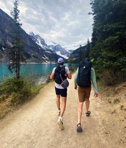Two runners jogging on a dirt trail along a turquoise alpine lake surrounded by pine forests and snow-capped mountains
