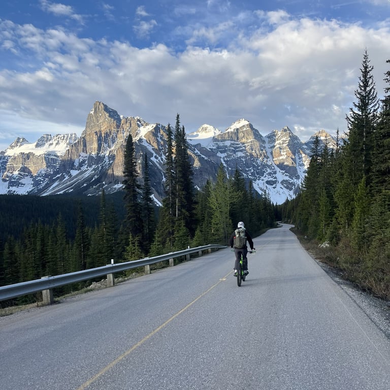 Cyclist riding on a mountain road with snow-capped peaks and evergreen forest in the background