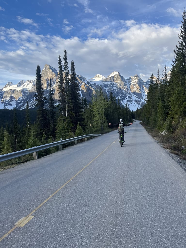 Cyclist on mountain road with snow-capped peaks and evergreen forest under blue sky