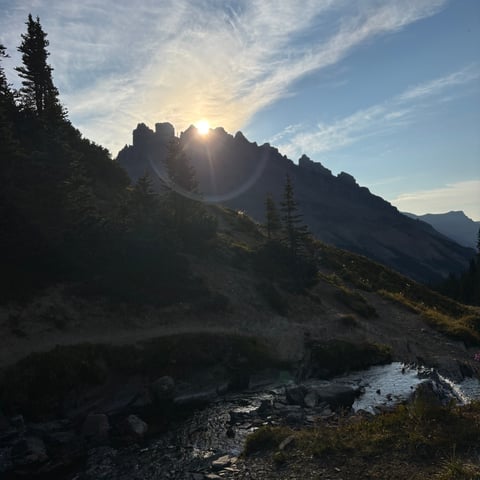 Mountain landscape at sunset with rocky peaks, pine trees, and a stream flowing through a misty valley