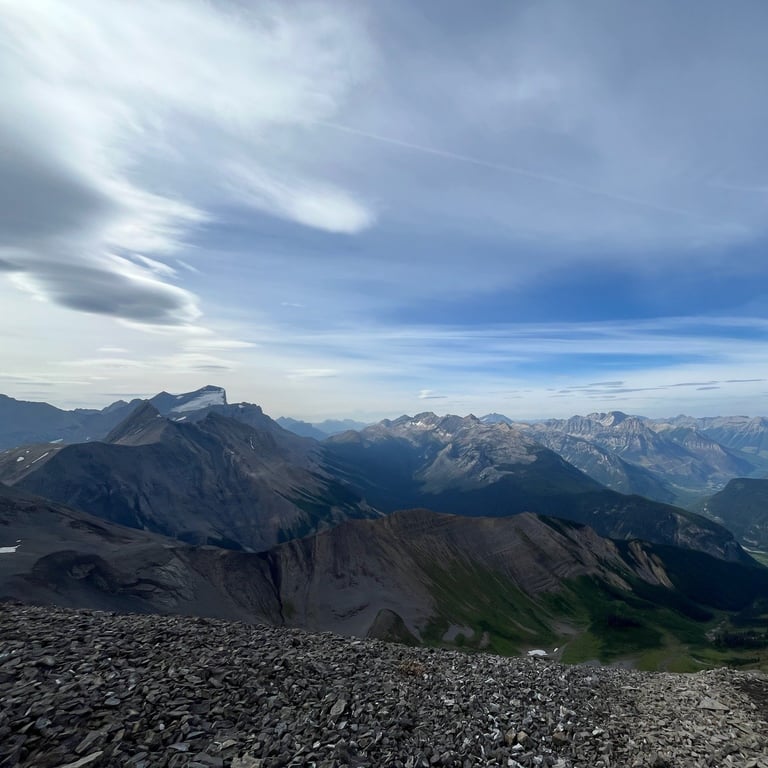 Panoramic mountain landscape with layered peaks, rocky foreground, and blue sky with wispy clouds