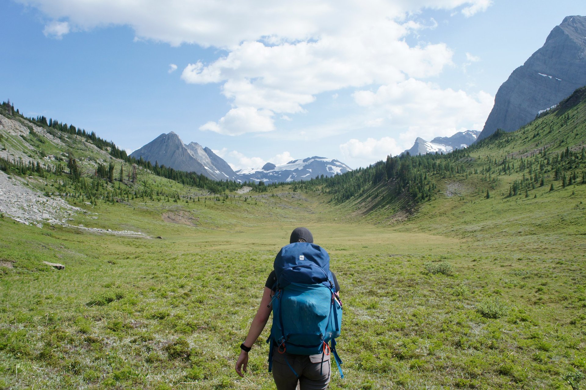 Hiker with blue backpack walking through green mountain valley surrounded by forested peaks and snow-capped mountains