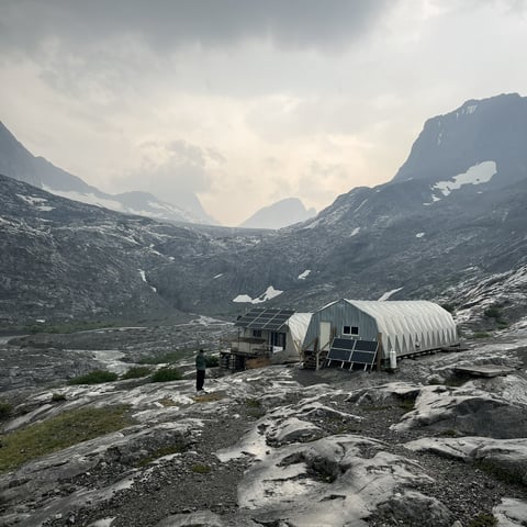 A metal tunnel shelter cabin sits on rocky terrain in a mountain valley with peaks and glaciers in the background under cloudy skies